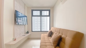 A modern living room with a beige sofa, a mounted TV, a window with blinds, and a small plant on a shelf. - Comfortable 1Br Apartment At Pollux Chadstone (Cikarang)