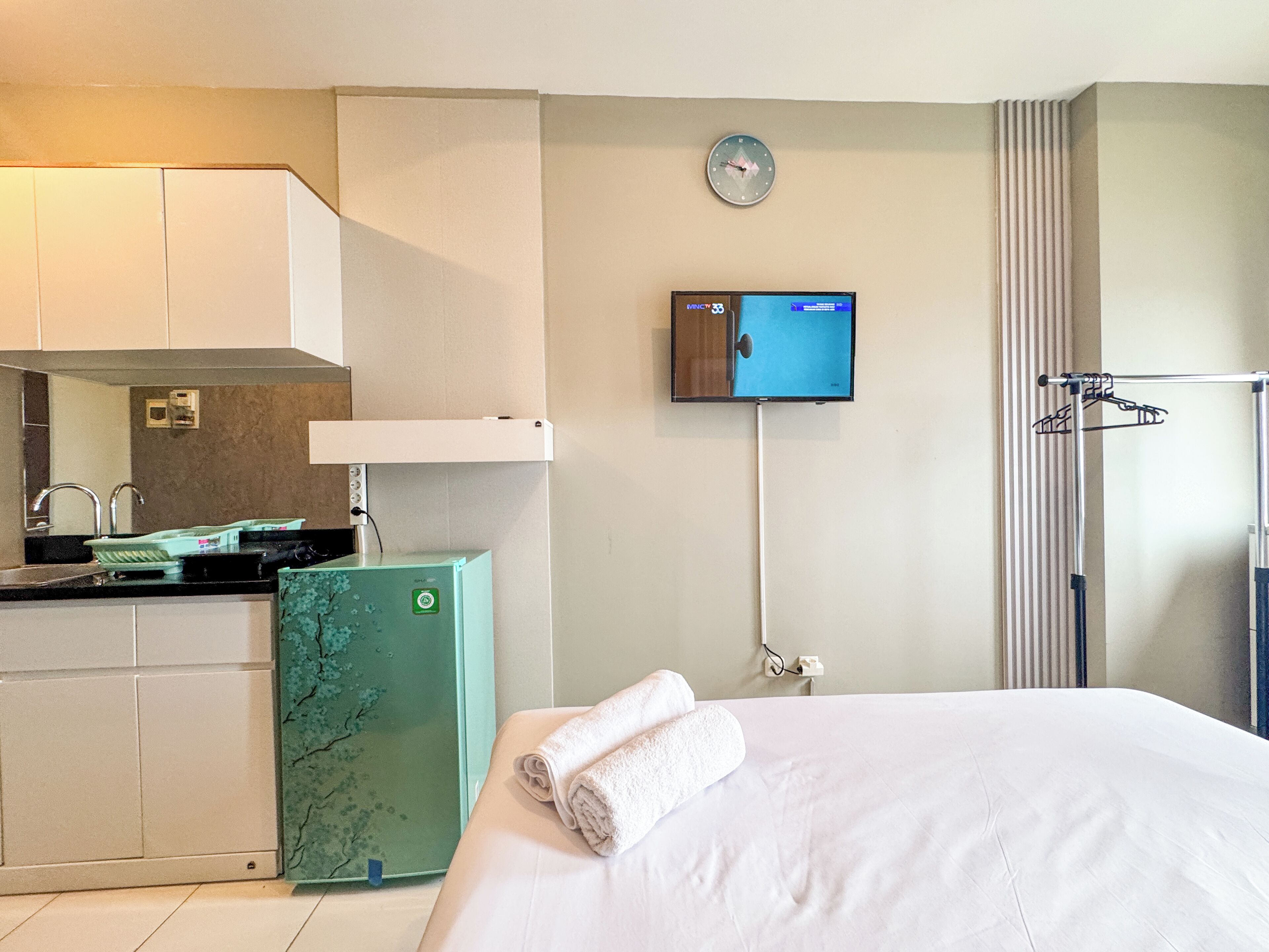 Habitación de hotel con cama, televisor montado en la pared, kitchenette pequeño y una toalla sobre la cama.