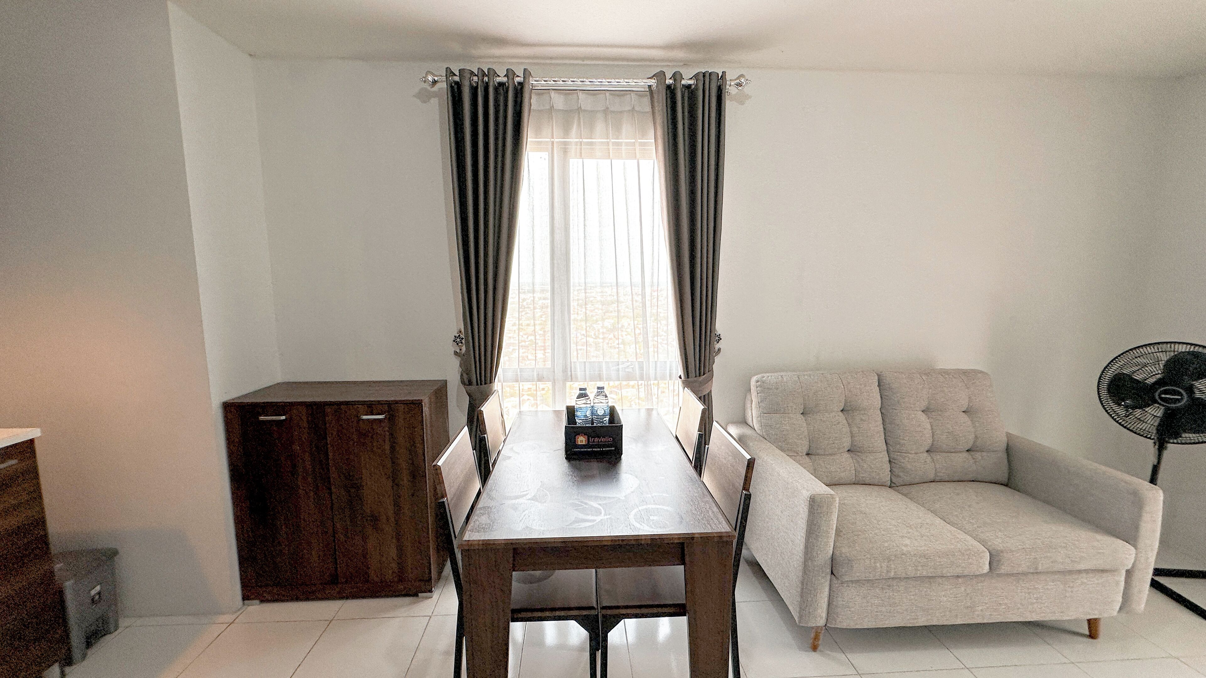 Une pièce comprenant une table à manger, un canapé, un meuble et un ventilateur. Il y a une fenêtre avec des rideaux et une vue sur la verdure à l’extérieur.