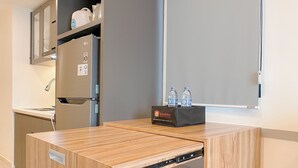 A compact kitchenette with a built-in refrigerator, a microwave, and a coffee maker. There is a small table with a water dispenser and a box labeled "Travelio." - Cozy And Modern 2Br At Meikarta Apartment (Cikarang)