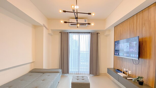 A modern hotel room with a sofa, ottoman, TV, and a wooden accent wall. - Cozy And Modern 2Br At Meikarta Apartment (Cikarang)