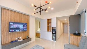 A modern living room with a flat-screen TV mounted on a wooden wall, a grey sofa, and a contemporary light fixture. - Cozy And Modern 2Br At Meikarta Apartment (Cikarang)