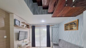 A modern living room with a sofa, a TV mounted on the wall, a wooden ceiling, and a patterned wall. - Elegant And Spacious 2Br Loft Green Park Yogyakarta Apartment (Depok)