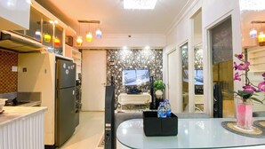 A modern kitchen with a refrigerator, oven, and microwave. There is a glass table with a black tray and a vase with flowers. A television is mounted on the wall, and there is a door leading to another room. - Good And Homey 1Br Apartment Mt Haryono Residence (Jakarta)
