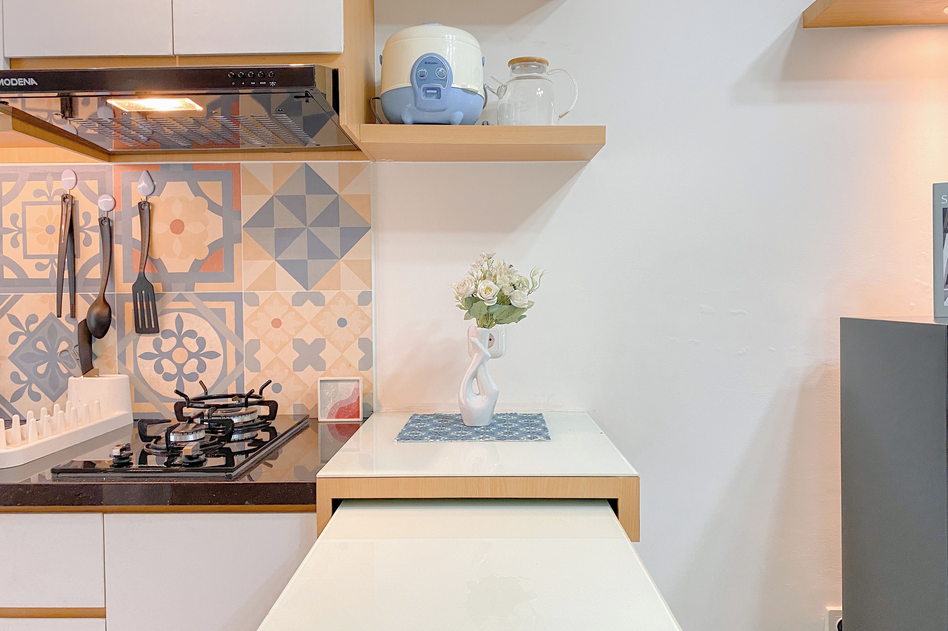 Una cocina moderna con estufa a gas, horno empotrado y campana extractora. Encima de la estufa hay un estante de madera con una tetera y un tarro. Un florero con flores se encuentra sobre una mesita frente a la estufa.