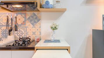 Una cocina moderna con estufa a gas, horno empotrado y campana extractora. Encima de la estufa hay un estante de madera con una tetera y un tarro. Un florero con flores se encuentra sobre una mesita frente a la estufa.