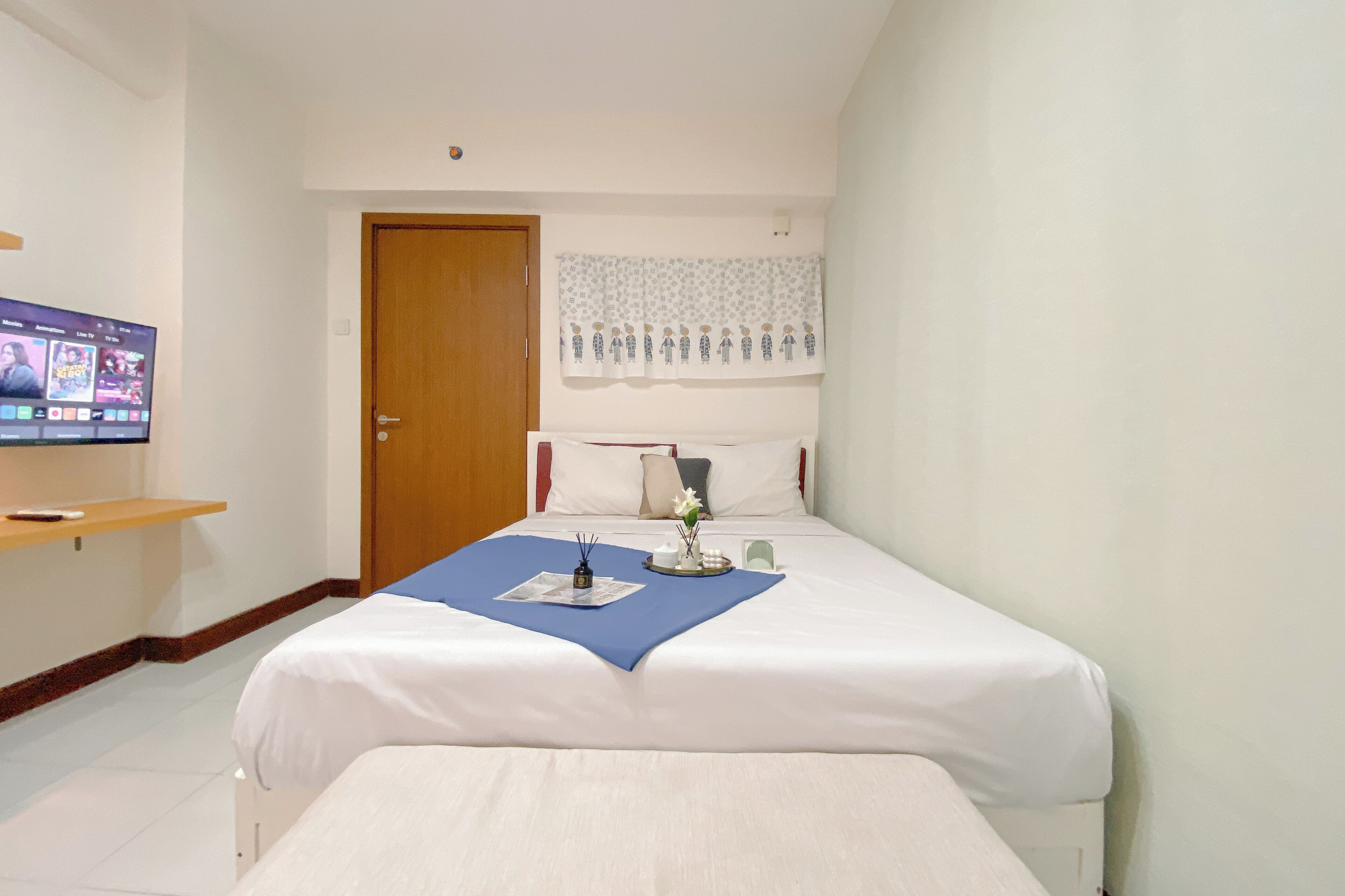 Quarto de hotel com uma cama, televisão na parede, porta de madeira e janela com cortina.