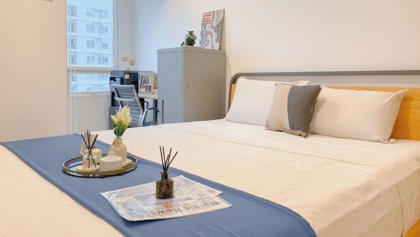 Ein ordentlich bezogenes Bett mit blauer Bettdecke, ein Tablett mit dekorativen Gegenständen und eine Zeitung. Außerdem ein grauer Nachttisch und ein Fenster mit Blick auf Gebäude.
