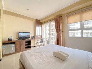 A hotel room with a bed, a TV, a desk with a computer, a mini-fridge, and a window with curtains. - Unique And Spacious Studio At Gateway Pasteur Apartment (Cimahi)
