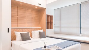 A modern bedroom with a wooden headboard, a large window with blinds, a bedside table with a magazine, and a bed with white bedding.