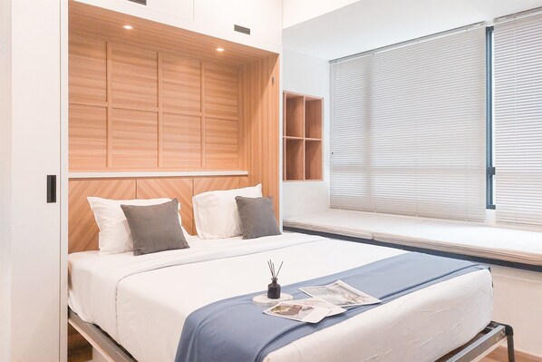 A modern bedroom with a wooden headboard, a large window with blinds, a bedside table with a magazine, and a bed with white bedding. - Good Choice And Homey Studio Apartment At Collins Boulevard (Tangerang)