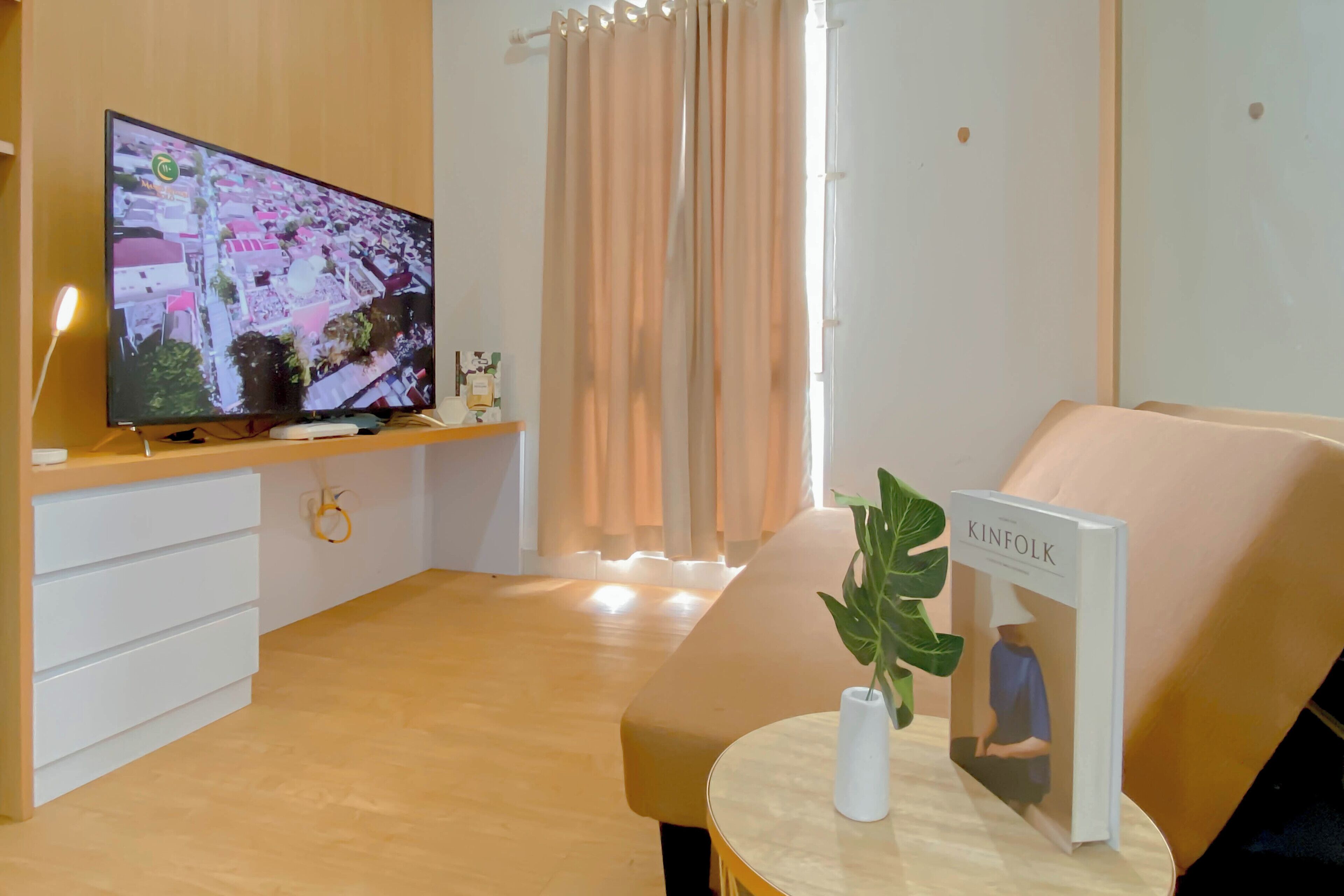 Egy modern nappali, melyben található egy televízió, egy kanapé, egy kis asztal, rajta egy növény és egy könyv, valamint egy ablak, melyen függöny van.