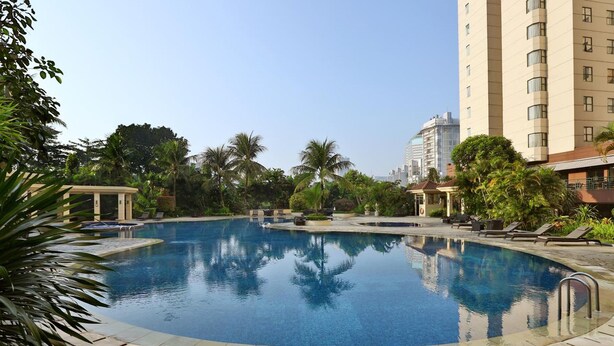 Una piscina amplia y cristalina rodeada de sillones de descanso y palmeras, con un edificio alto al fondo.