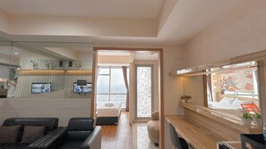 A modern hotel room with a large bed, a desk, a chair, a TV, and a view of the city through the window. - Comfortable Living And Best Location 1Br At Grand Sungkono Lagoon Apartment (Surabaya)
