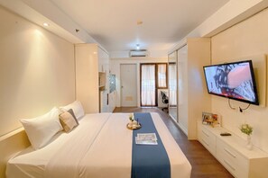 A hotel room with a bed, a TV mounted on the wall, a bedside table with a vase, and a door leading to another room. - Cozy Studio At Metropark Condominium Apartment (Cikarang)