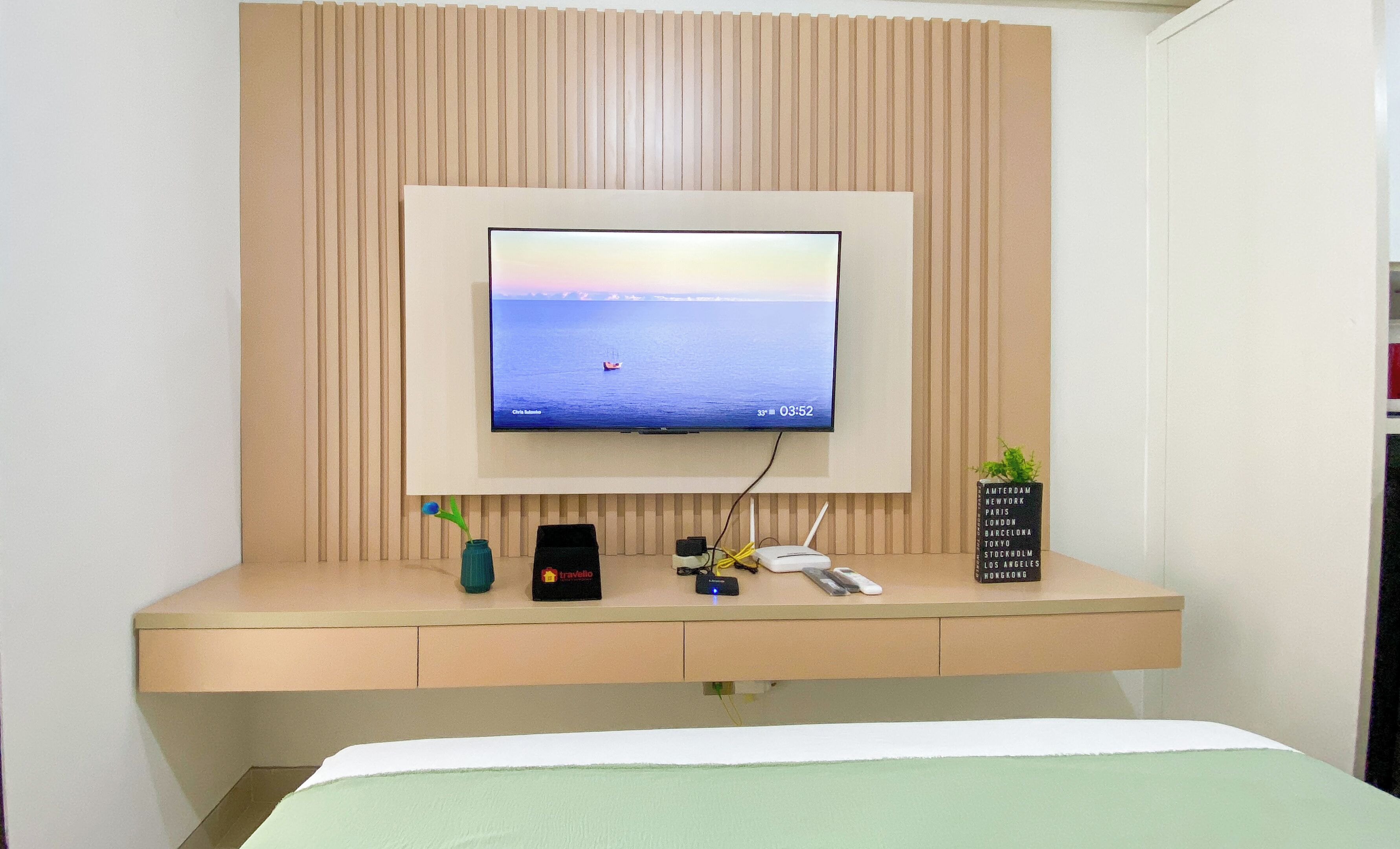 A bedroom with a bed, a TV mounted on a wall, a desk with a laptop, a phone, and a small plant.
