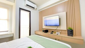 A hotel room with a bed, a TV mounted on a wooden panel wall, a window, and an air conditioning unit. - Best Value Studio Room Transpark Cibubur Apartment (Depok)