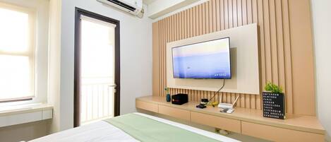 Una habitación de hotel con cama, televisor montado en una pared de paneles de madera, ventana y equipo de aire acondicionado.