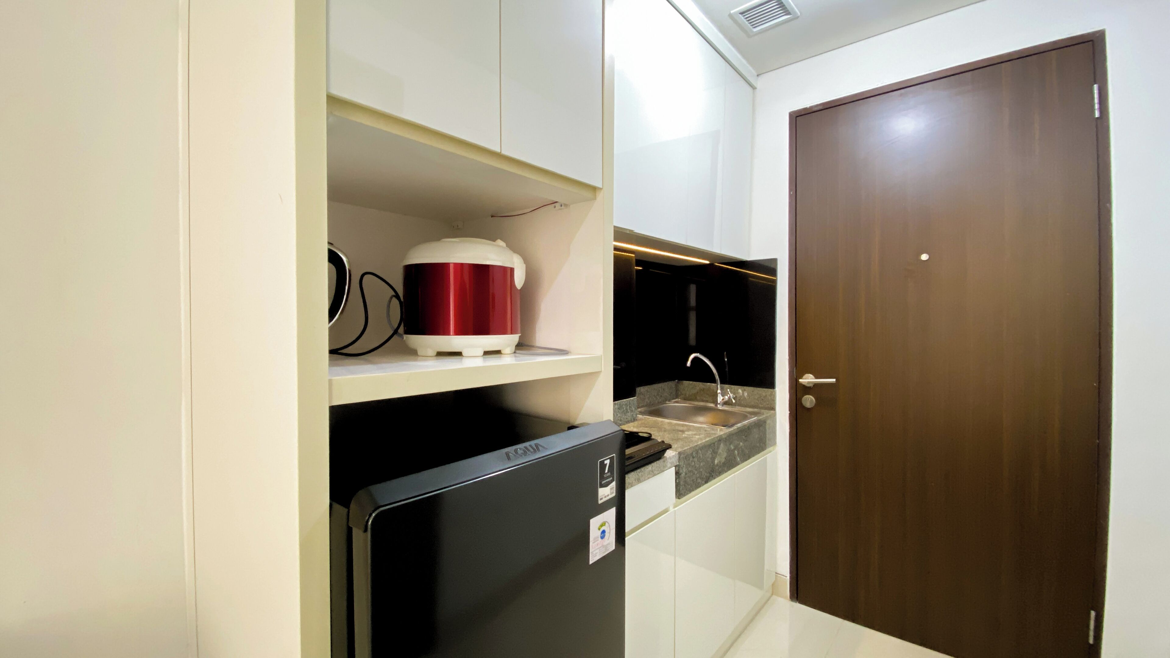 Una cocina compacta con armarios blancos, un refrigerador negro, una tetera eléctrica roja y blanca, y una puerta de madera.