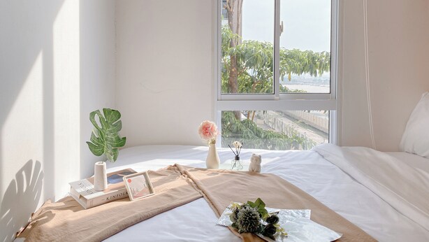 Un lit bien fait, avec vue sur la verdure à l’extérieur, une plante en pot, un vase rempli de fleurs et quelques livres posés sur le lit.