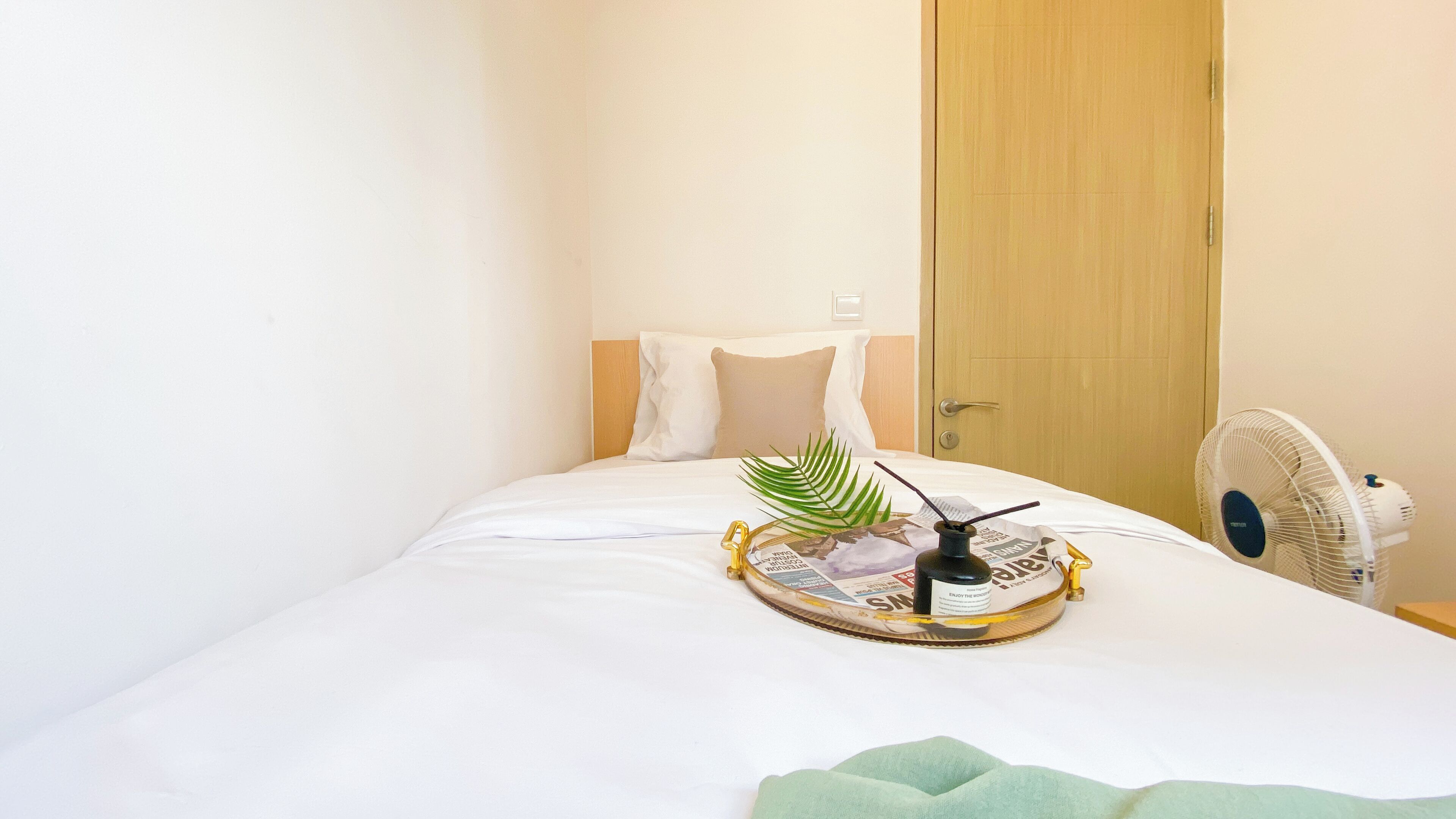 A neatly made bed with white linens, a small decorative plant, and a fan on the side.