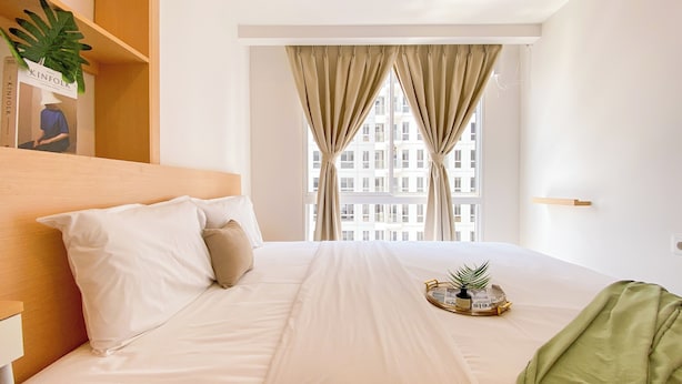 A hotel room with a bed, a nightstand, a window with curtains, and a plant on the windowsill. - Comfortable And Simply 2Br At Tokyo Riverside Pik 2 Apartment (Teluknaga)