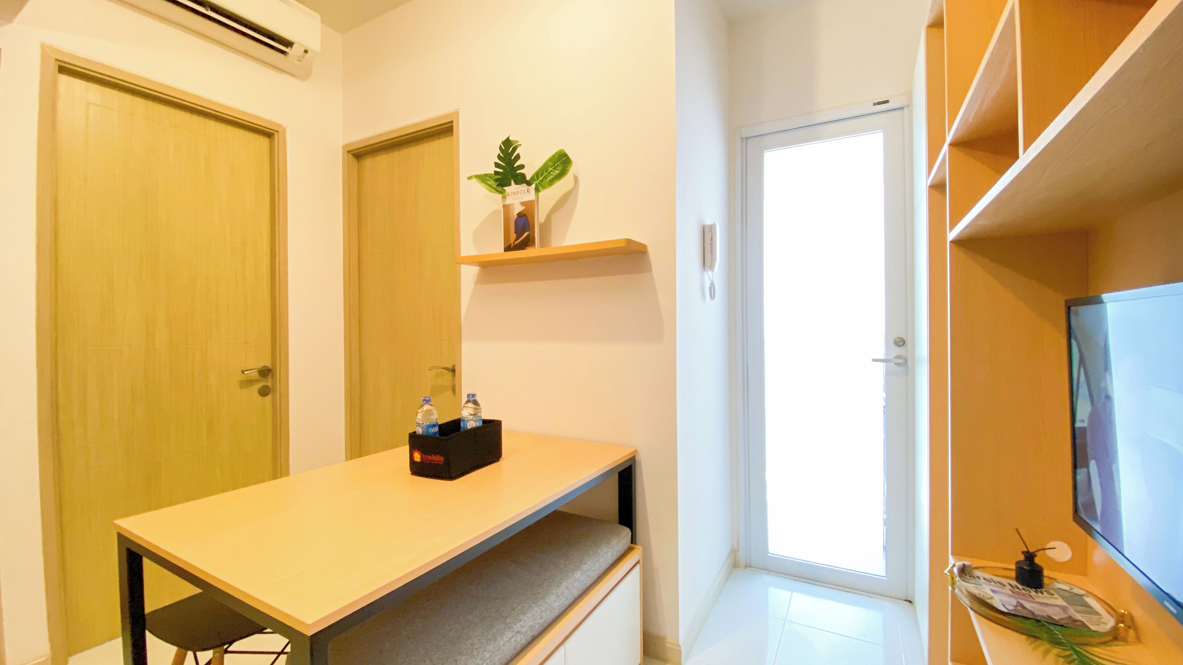 A compact room with a wooden desk, a small shelf with a plant, a television, and a door leading to another room.
