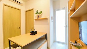 A compact room with a wooden desk, a small shelf with a plant, a television, and a door leading to another room. - Comfortable And Simply 2Br At Tokyo Riverside Pik 2 Apartment (Teluknaga)