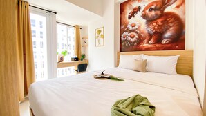 Habitación de hotel con una cama grande, un escritorio con silla, una ventana con cortinas y un cuadro con un conejo y flores en la pared.