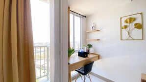 A modern room with a wooden desk, a black chair, a window with a view of buildings, and a framed artwork on the wall. - Modern And Tranquil Studio At Apartment Tokyo Riverside Pik 2 (Teluknaga)