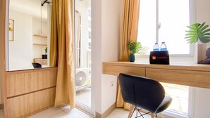 Une chambre d’hôtel moderne avec un bureau en bois, une chaise noire, un grand miroir et une fenêtre avec des rideaux. On trouve deux bouteilles d’eau et une plante sur le bureau.