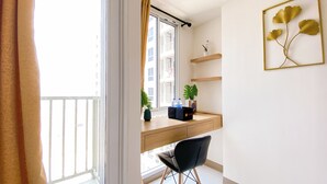 Une chambre moderne équipée d’un bureau, d’une chaise, de plantes en pot et offrant une vue sur un immeuble par la fenêtre.