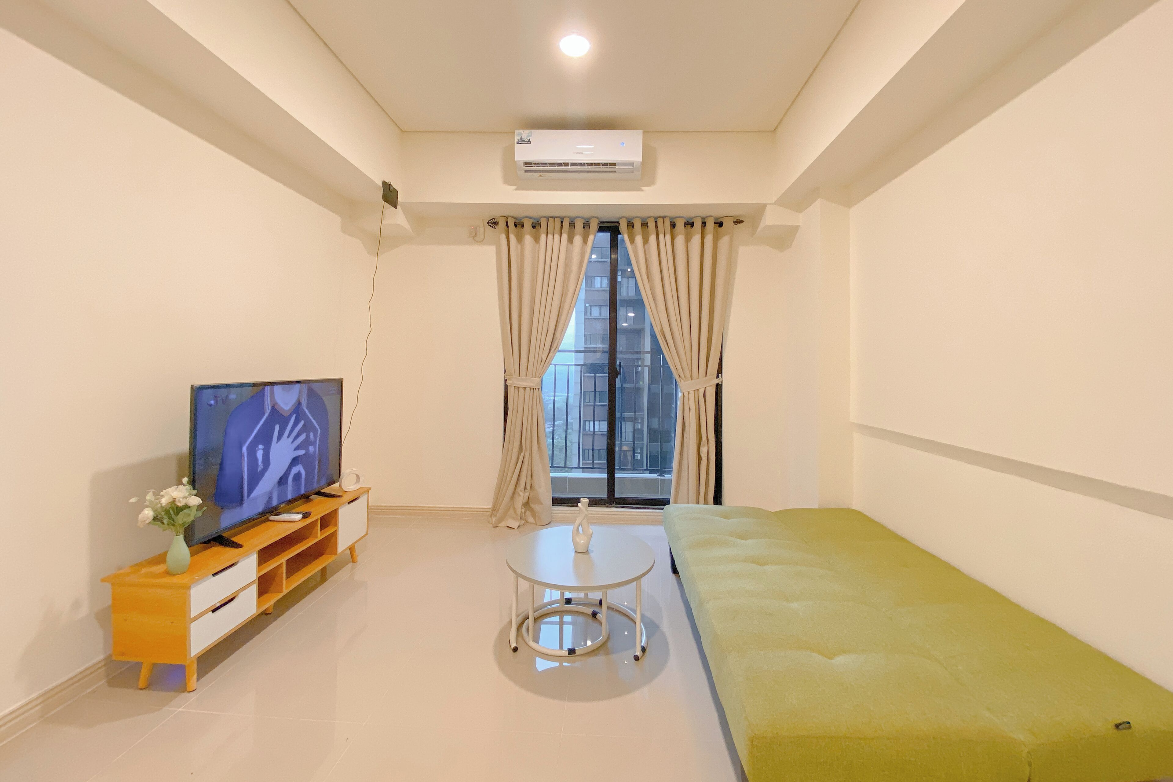 Quarto moderno com televisão de tela plana sobre um móvel de madeira, uma pequena mesa branca, um sofá verde e uma janela com cortinas.