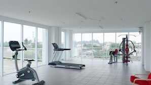 A gym with a treadmill, elliptical, and weight machines, large windows, and a red chair. - Homey And Cozy Living Studio At Poris 88 Apartment (Tangerang)