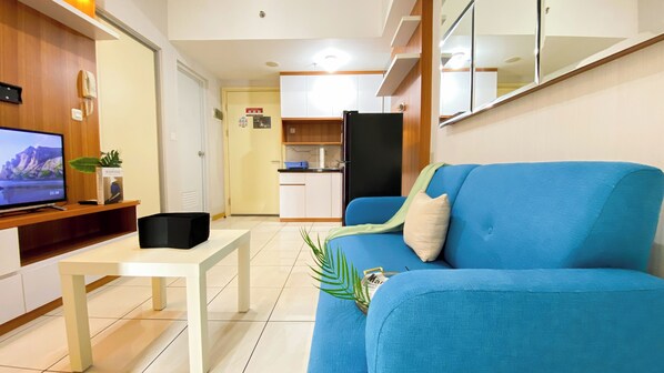 A living room with a blue sofa, a TV on a wooden stand, a small white table, and a kitchen in the background. - Scenic 2Br Apartment At M-Town Residence (Gading Serpong)