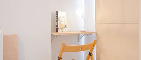 A small, compact room with a wooden chair, a shelf with a book and a perfume bottle, and a bedside table with a lamp.