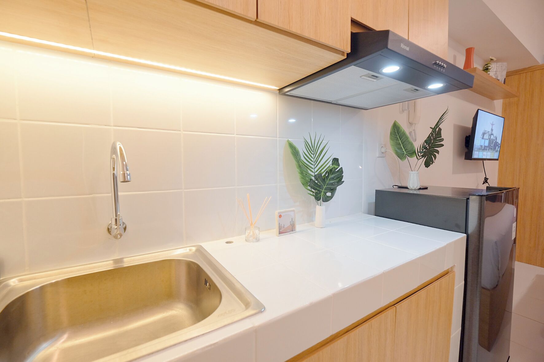 Una cocina moderna con un lavamanos de acero inoxidable, encimera blanca, armarios de madera, una campana extractora negra y una pequeña planta en maceta.