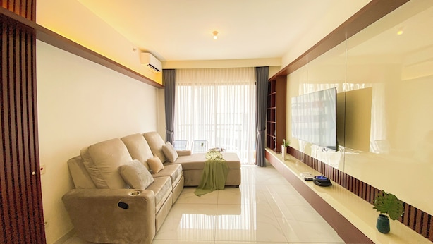 Una sala de estar moderna con un sofá beige, un televisor de pantalla plana en la pared y un ventanal con cortinas traslúcidas.