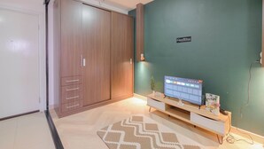 A modern room with a wooden wardrobe, a flat-screen TV on a wooden stand, and a green accent wall. - Comfort And Big 1Br At Green Pramuka City Apartment (Jakarta)