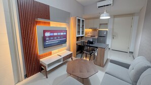 A modern living room with a flat-screen TV mounted on a wooden panel, a sofa, a coffee table, and a dining area with a small table and chairs. - Minimalist 2Br And Well Design Room At Benson Supermall Mansion Apartment (Surabaya)