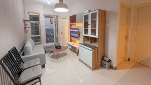 A modern living room with a sofa, coffee table, and TV. There's a kitchen area with cabinets and a trash bin. - Minimalist 2Br And Well Design Room At Benson Supermall Mansion Apartment (Surabaya)