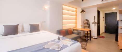 A modern hotel room with a large bed, a desk, and a chair. There is a small table with a lamp, and a door leading to another room.