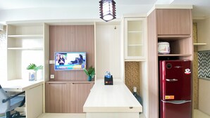 A compact kitchen with a TV mounted on the wall, a small table with a plant, a refrigerator, and a cabinet with a water dispenser. - Great City View Studio At Tamansari The Hive Apartment (Jakarta)