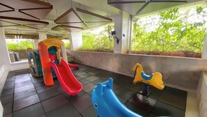 A children's play area with slides and swings, large windows with greenery outside, and a tiled floor.