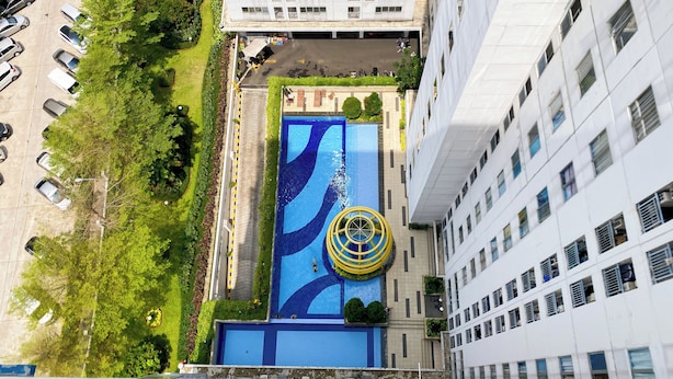 An aerial view of a building with a swimming pool, surrounded by greenery and adjacent to a parking area.