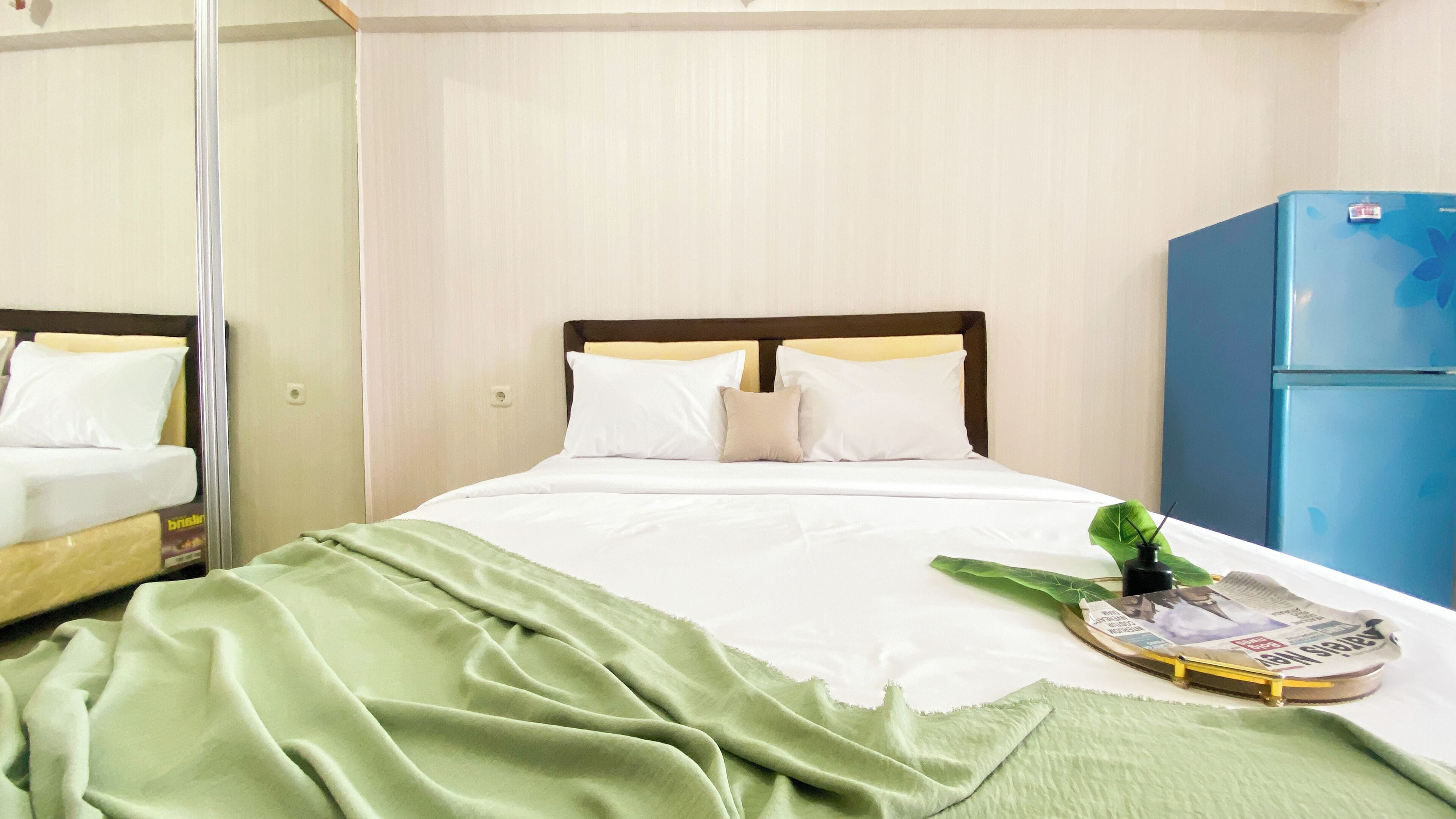 A hotel room with a bed, a green blanket, a small plant, and a blue refrigerator.