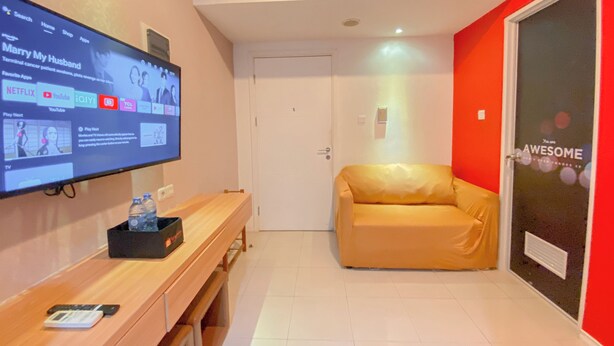 A modern living room with a flat-screen TV mounted on the wall displaying a YouTube video. There is a wooden bench with a smartphone and a water bottle on it. A beige sofa is positioned against a red wall with a door that has a 'AWESOME' sign. - Best Deal 1Br Apartment Parahyangan Residence (Bandung)