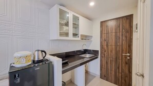 A compact kitchen with white cabinetry, a black countertop, a wooden door, and a refrigerator with a kettle on top. - Comfortable Studio At Altuz Seturan Yogyakarta Apartment (Depok)