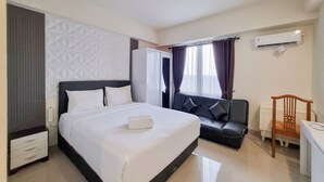 A hotel room with a bed, a desk, a chair, a wardrobe, and an air conditioning unit. - Comfortable Studio At Altuz Seturan Yogyakarta Apartment (Depok)