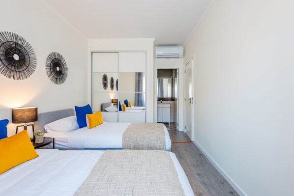 Habitación de hotel con dos camas, una mesita de noche con lámpara, un espejo y una kitchenette al fondo.